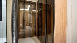 Quarto de hotel moderno, com divisória de vidro, piso de madeira e banheiro visível com chuveiro e vaso sanitário.