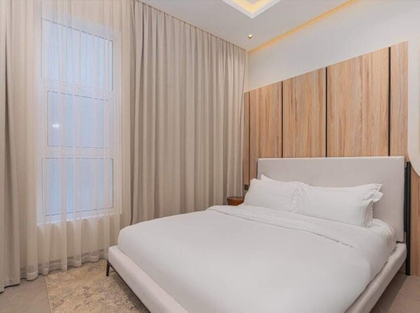 Une chambre à coucher moderne avec un grand lit, une tête de lit en bois et une fenêtre ornée de rideaux légers.