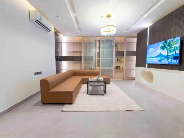 A modern living room with a sectional sofa, a coffee table, a TV mounted on the wall, and a decorative chandelier. - Trendy Apartment in Alwadi lX (Riyadh)