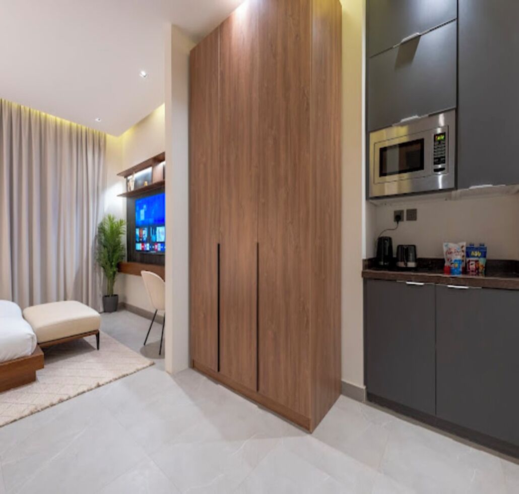 A modern hotel room with a bed, a seating area, a kitchenette, and a large wardrobe.