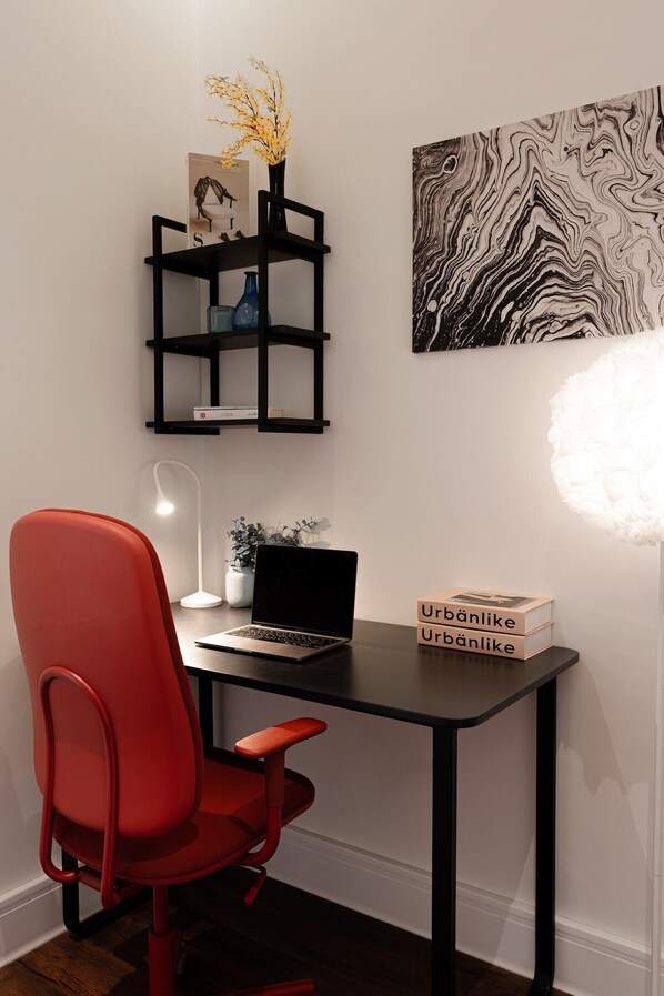 A modern workspace with a red chair, a black desk, a laptop, a black shelving unit with decorative items, and a wall-mounted abstract art piece.