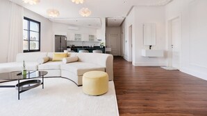 A spacious living room with a white sofa, a round coffee table, and a yellow ottoman. There is a kitchen in the background with white cabinets and a refrigerator. - Luxury Apartment in Alnada (Riyadh)