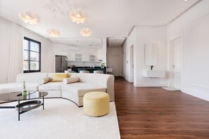Ein geräumiges Wohnzimmer mit einem weißen Sofa, einem runden Couchtisch und einem gelben Hocker. Im Hintergrund befindet sich eine Küche mit weißen Schränken und einem Kühlschrank.