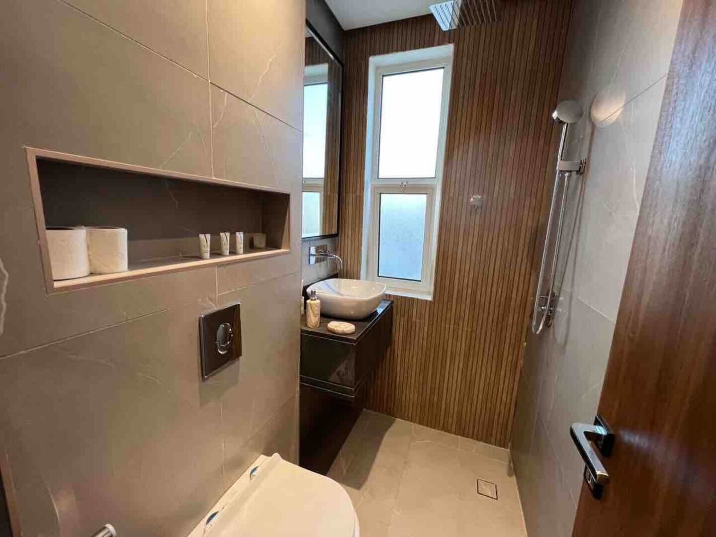 Un baño moderno con inodoro, lavabo y ducha. Cuenta con una ventana que proporciona luz natural y un estante con artículos de aseo.