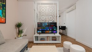 A modern living room with a flat-screen TV, a white entertainment unit, a sofa, two ottomans, and a decorative screen. - One Bedroom Trendy Apartment in Almughrizat (Riyadh)