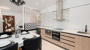 A modern kitchen with a dining area, featuring a built-in oven, microwave, and a large window with blinds. - One Bedroom Trendy Apartment in Almughrizat (Riyadh)