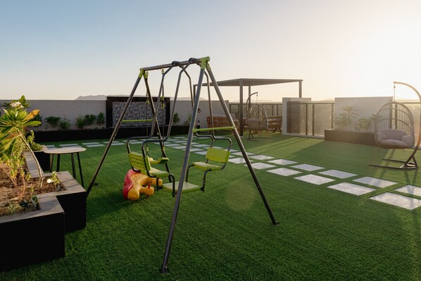 A rooftop terrace with artificial grass, a swing set, a hanging chair, and a seating area with a table and chairs.