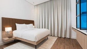 A modern hotel room with a wooden headboard, a white bed, a nightstand with a lamp, a large window with curtains, and a wooden floor. - Modern Comfort, Dedicated Desk, Free Parking (Riyadh)