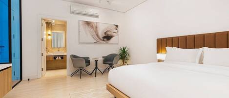 A modern hotel room with a large bed, a small desk with two chairs, a bathroom with a sink and mirror, and a large window.