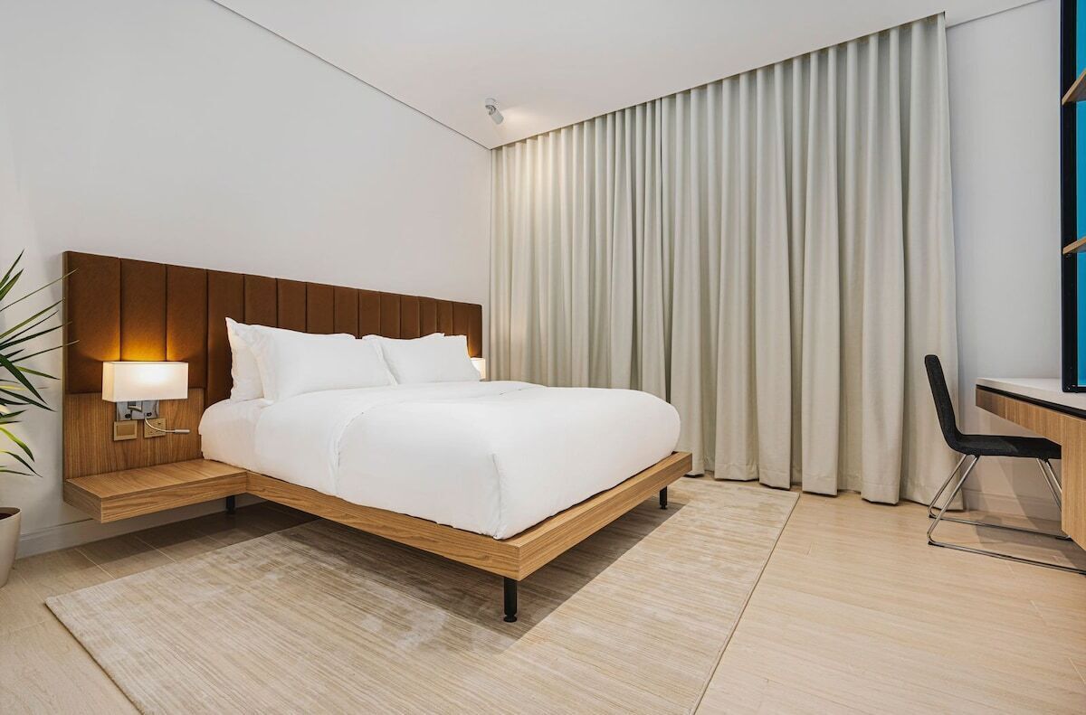 Una habitación de hotel moderna con una cama de madera, un escritorio y un amplio ventanal con cortinas.