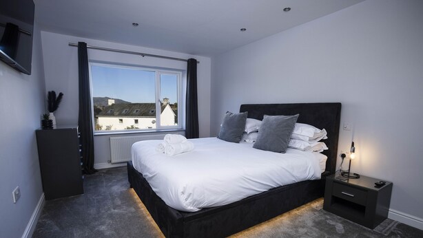 Un dormitorio moderno con una cama grande, una mesita de noche con lámpara y vistas a edificios a través de una ventana.