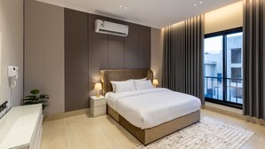 A modern bedroom with a large bed, bedside lamps, an air conditioning unit, a small plant, and a view of the outside through a window.