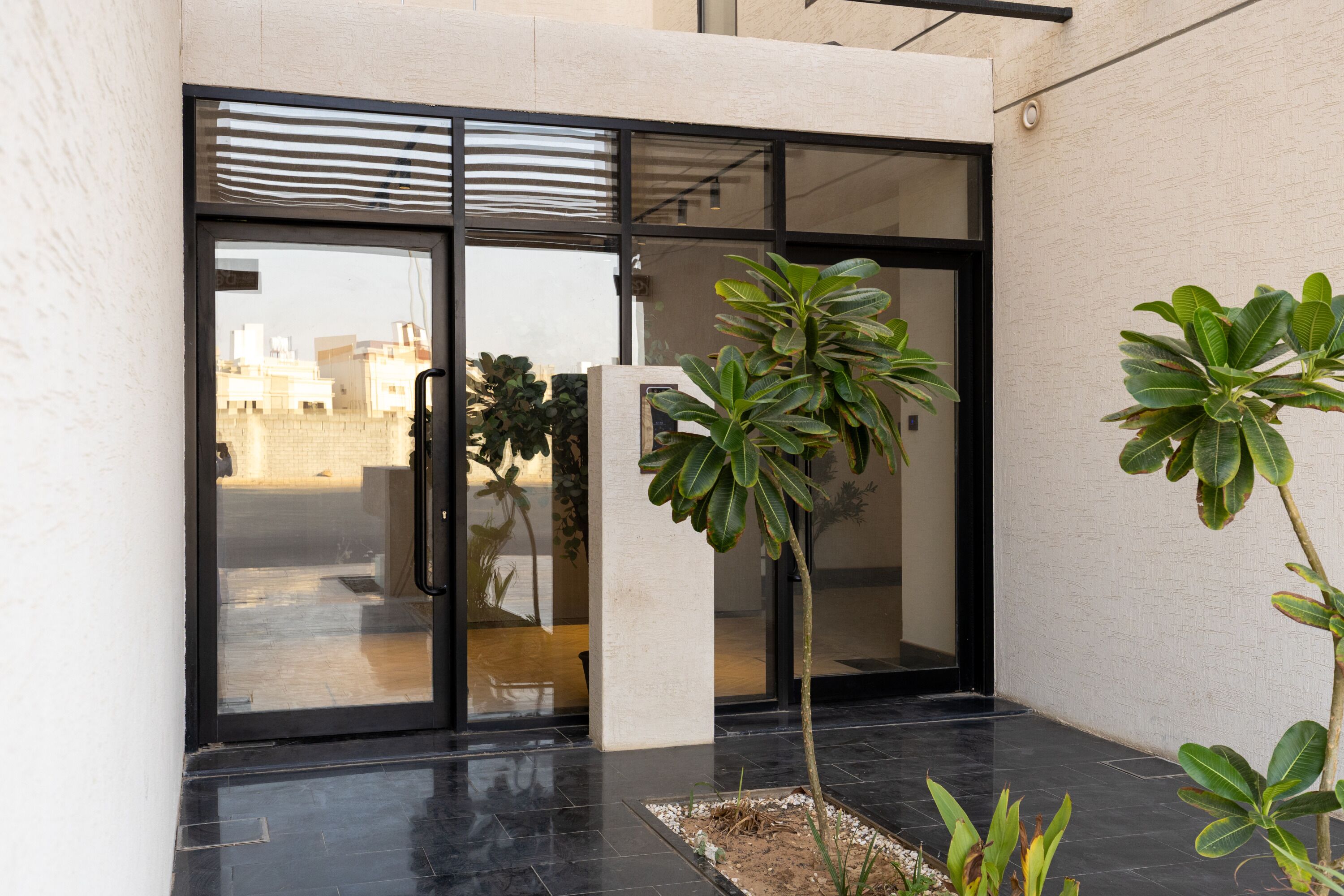 A modern building entrance with glass doors, a concrete pillar, and a small garden area with plants.