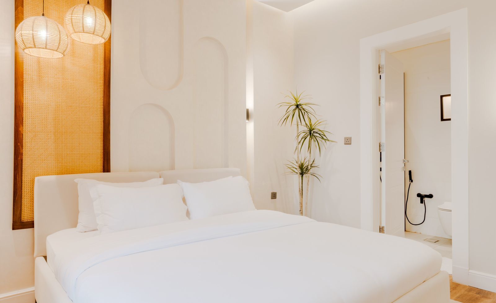 Une chambre moderne avec un grand lit, deux lampes suspendues, une tête de lit en bois et une porte donnant sur une salle de bain.