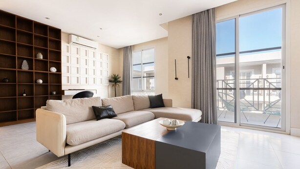 A modern living room with a sectional sofa, a coffee table, and a large window with a view of the balcony. - Exclusive Stay With Cinema, Wifi, Workspace (Riyadh)