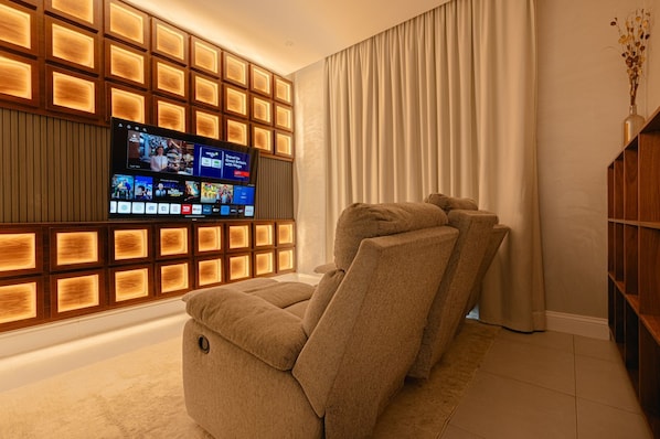 Una sala de estar moderna con un televisor de pantalla plana grande montado en una pared de paneles de madera, un sofá color beige y una estantería de madera.