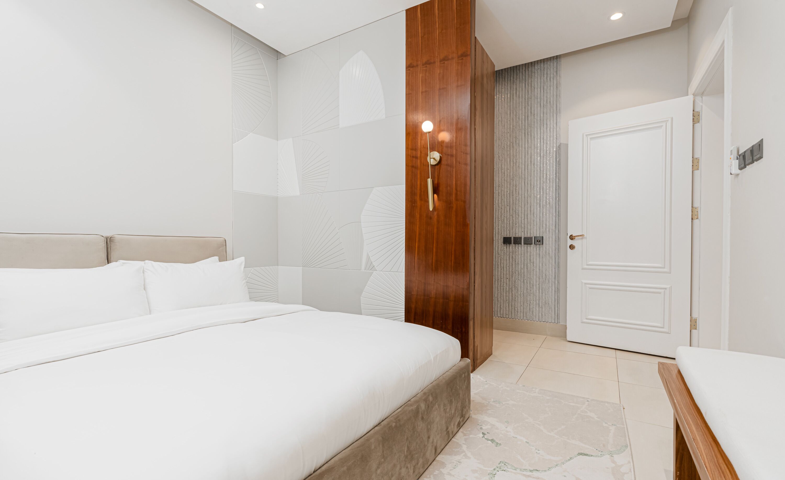 A modern hotel room with a large bed, a wooden wardrobe, a white door, and a textured wall.