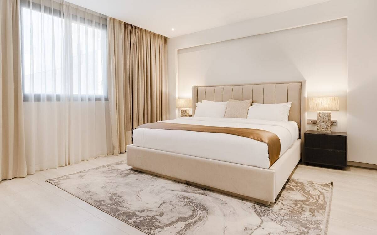 Ein modernes Hotelzimmer mit einem großen Bett, zwei Nachttischen mit Lampen, einem großen Fenster mit Vorhängen und einem gemusterten Teppich.