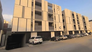 Un immeuble à étages doté d’une architecture moderne, avec une série de balcons et un espace de stationnement où sont garées plusieurs voitures.
