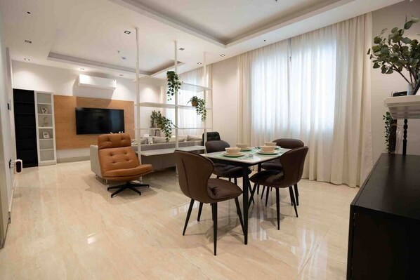 Un comedor moderno con una mesa dispuesta para cuatro personas, rodeada de sillas. Hay un televisor montado en la pared, un sofá y una planta en el alféizar de la ventana.