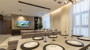 Uma sala de estar moderna com uma grande TV de tela plana, um sofá, uma mesa de jantar posta com pratos e velas, e uma janela grande com cortinas finas.