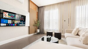 Una sala moderna con un televisor de pantalla plana montado en la pared que muestra varios canales y aplicaciones. Hay un sofá color beige, una mesita de centro y una planta en maceta.