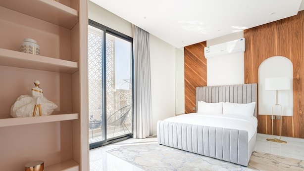 Una habitación de hotel moderna con una cama grande, un panel de madera decorativo, una puerta de vidrio que da a un balcón y una estantería empotrada con objetos decorativos.