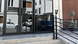 Moderne bygningseingang med glasfacade, trappe op til indgangen og en parkeringsplads med flere biler.