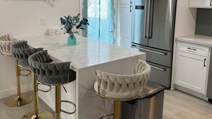 Une cuisine moderne dotée d’un îlot central, d’un réfrigérateur en acier inoxydable, de meubles blancs et de tabourets de bar aux pieds dorés.