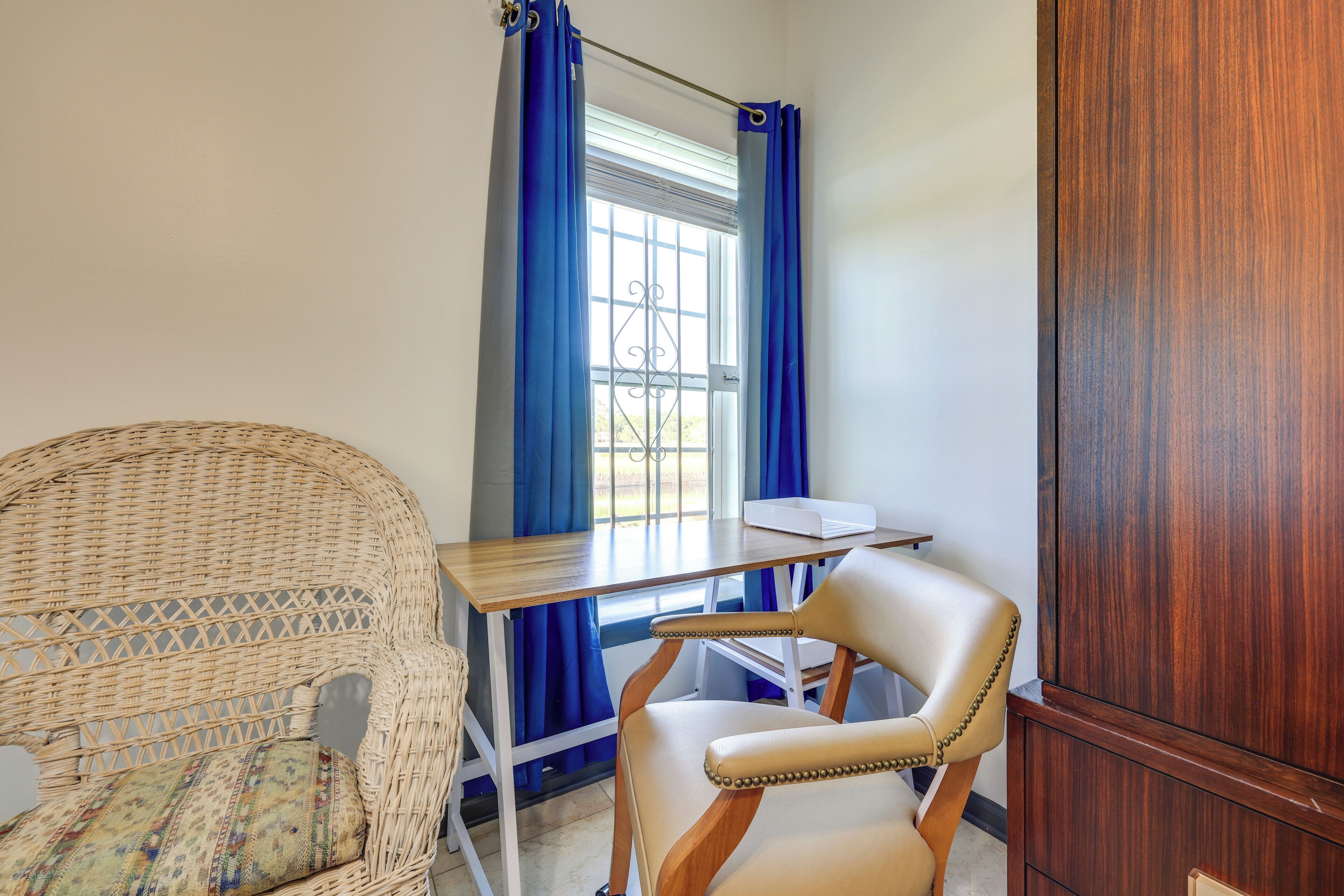 A room with a wicker chair, a wooden desk, a leather chair, and a window with blue curtains.