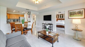 A modern living room with a sofa, coffee table, and dining area. There is a kitchen in the background with wooden cabinets and a refrigerator. A flat-screen TV is mounted on the wall, and a painting is visible on the wall. - Golf Views: Kissimmee Condo on Reunion Course (Kissimmee)
