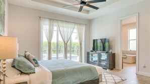 Un dormitorio con cama, ventilador de techo, televisor y baño visible a través de una puerta abierta.