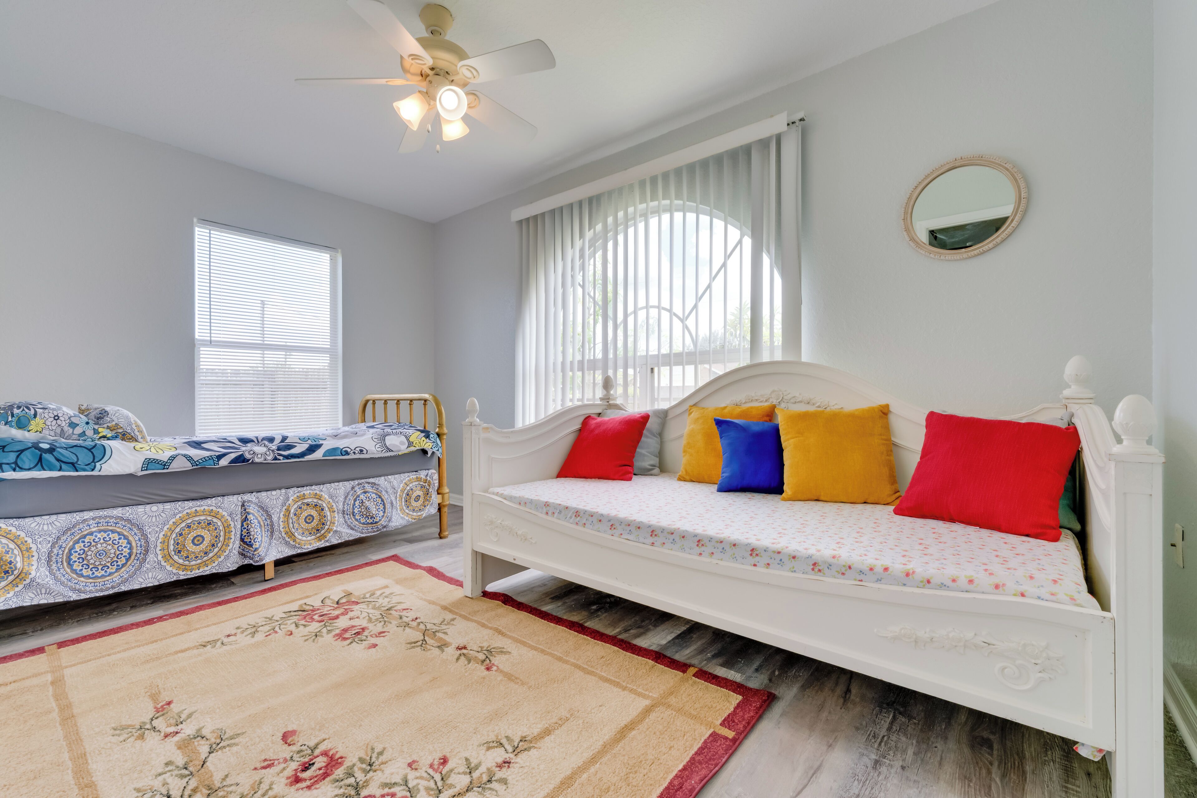 A bedroom with a white bed, colorful pillows, a ceiling fan, and a large window with curtains.