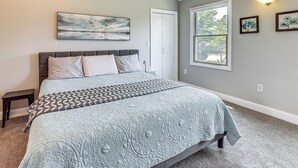 Um quarto com cama, mesa de cabeceira, janela e quadros nas paredes.