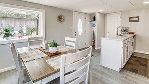 Uma sala de jantar com mesa de madeira, cadeiras brancas e área de cozinha com armários brancos e pia.