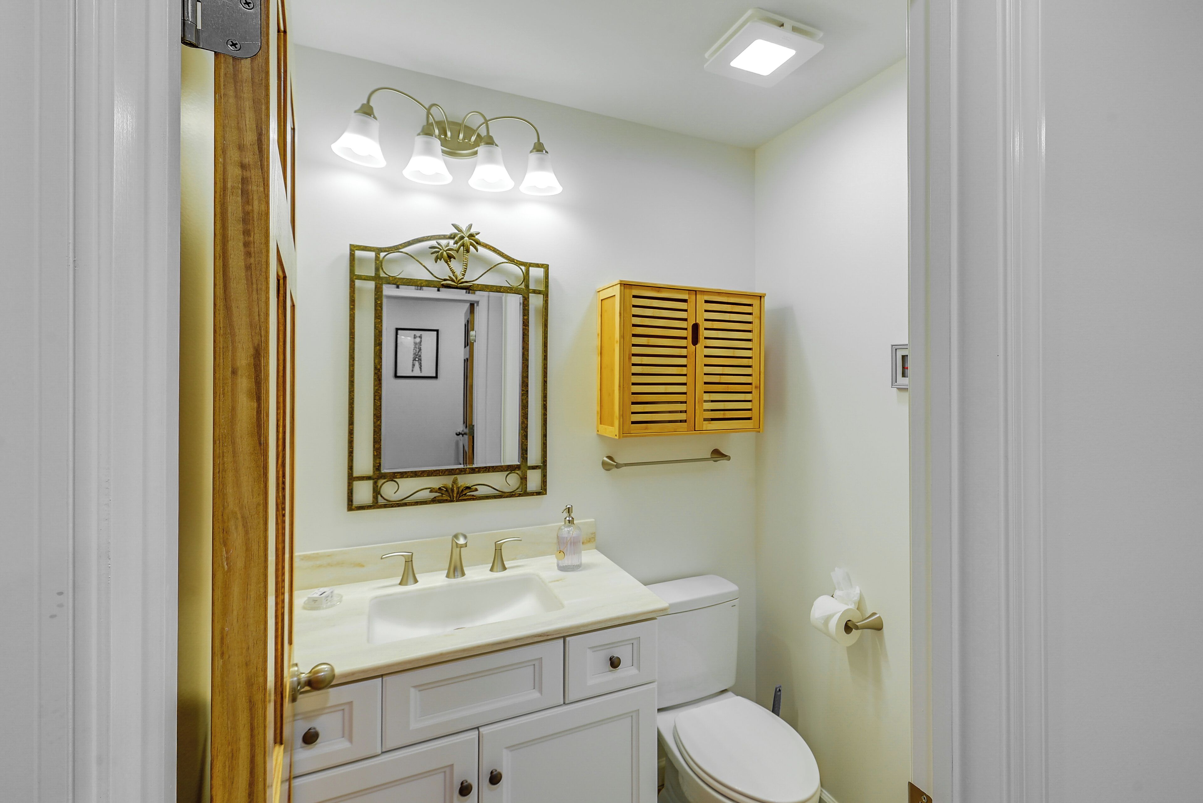 A bathroom with a white sink, vanity, and toilet. There is a mirror with a decorative frame, a wooden cabinet, and a light fixture above the sink.