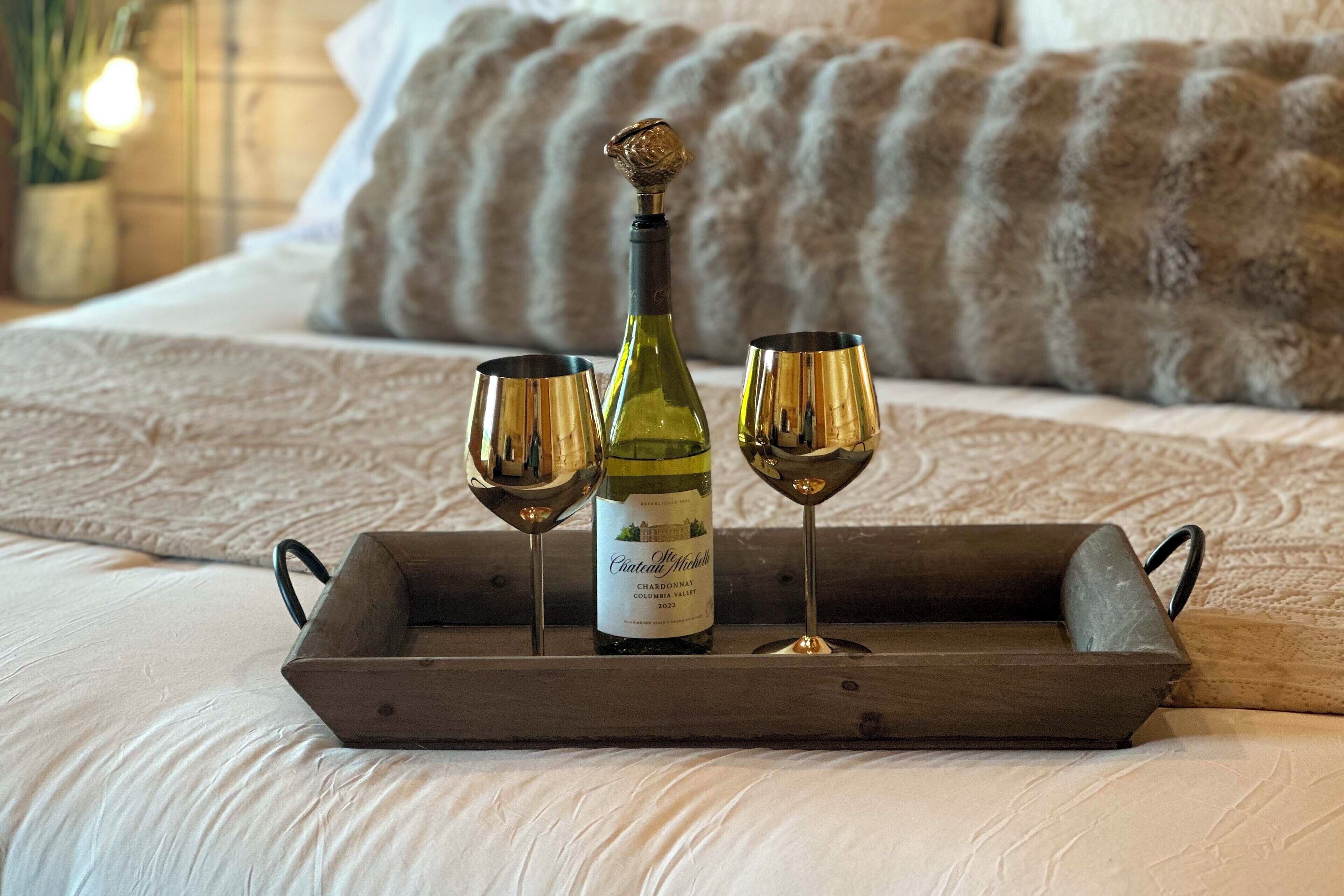 A wooden tray with a bottle of Chateau McConnell Chardonnay 2022 and two glasses on a bed with a textured blanket and pillows.