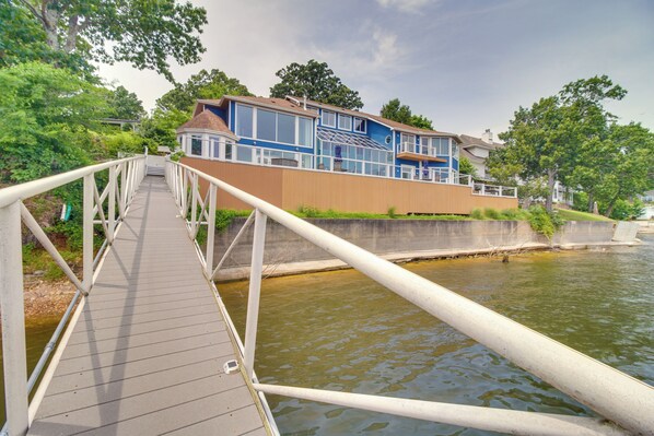 A wooden bridge leading to a house with a large window and a balcony.