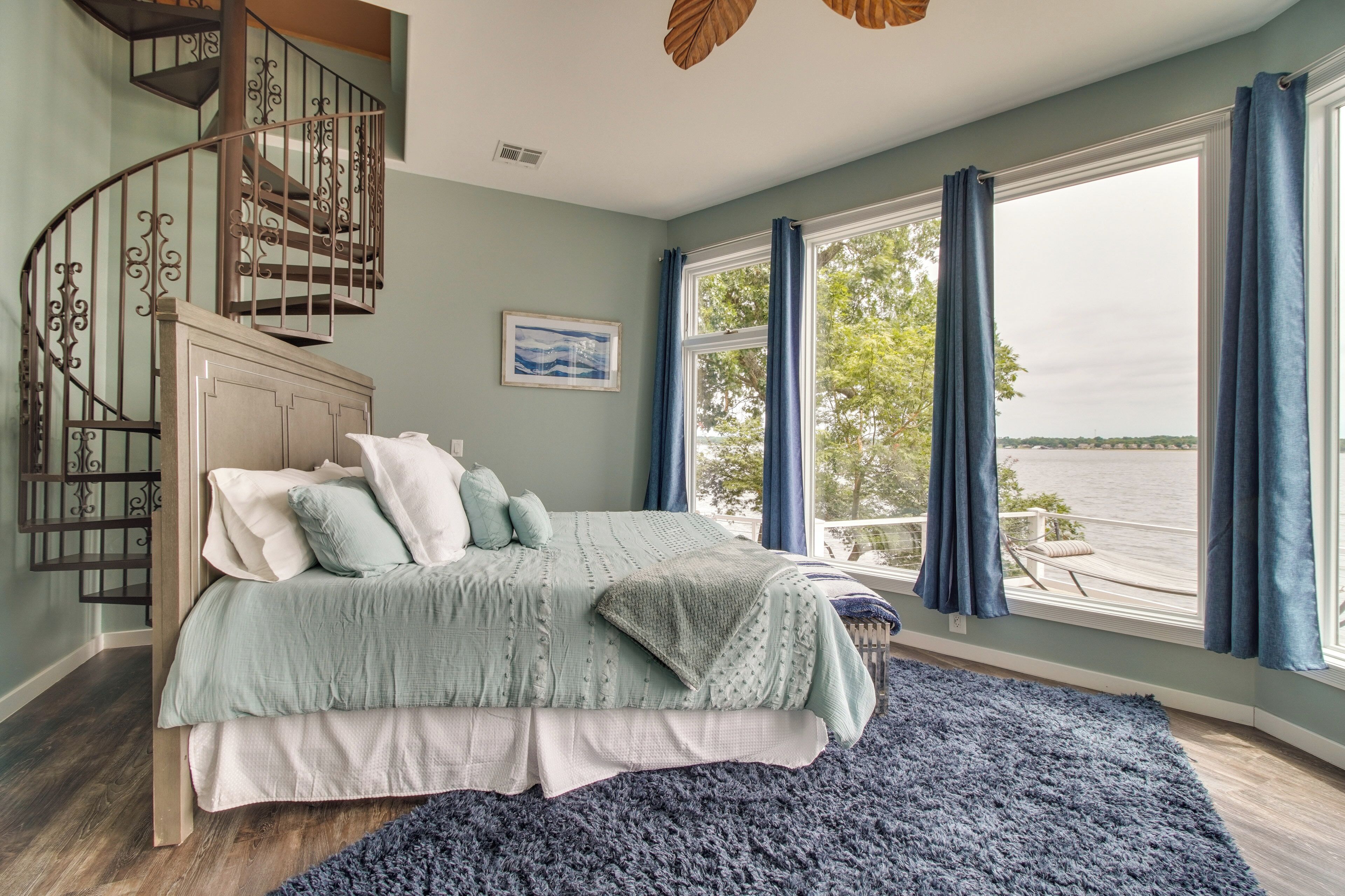 A bedroom with a bed, a view of the water, a staircase, and a window with curtains.