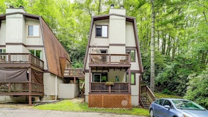 Two multi-story buildings with balconies, a car parked in front, and a forested background.
