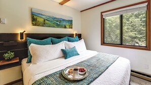 A neatly made bed with a tray of breakfast items, a painting of a landscape on the wall, and a window with a view of trees.