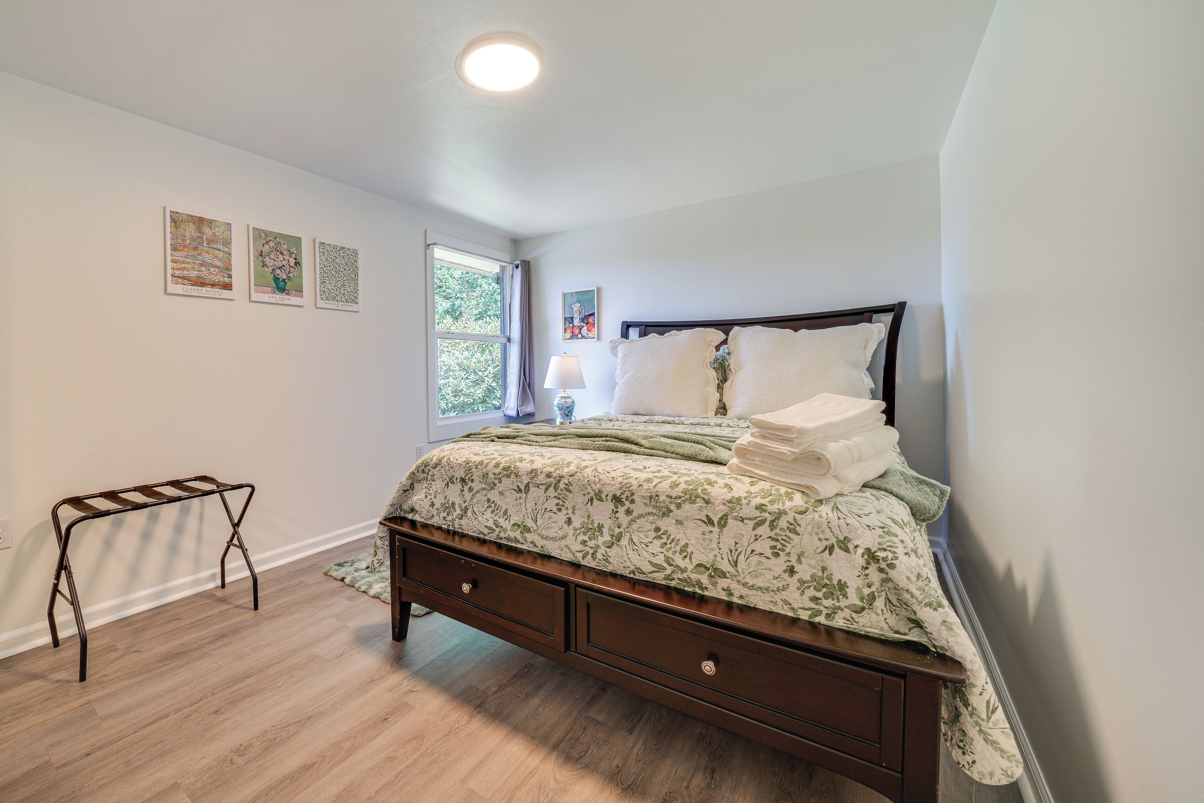 A bedroom with a bed, a nightstand, a window, and artwork on the wall.