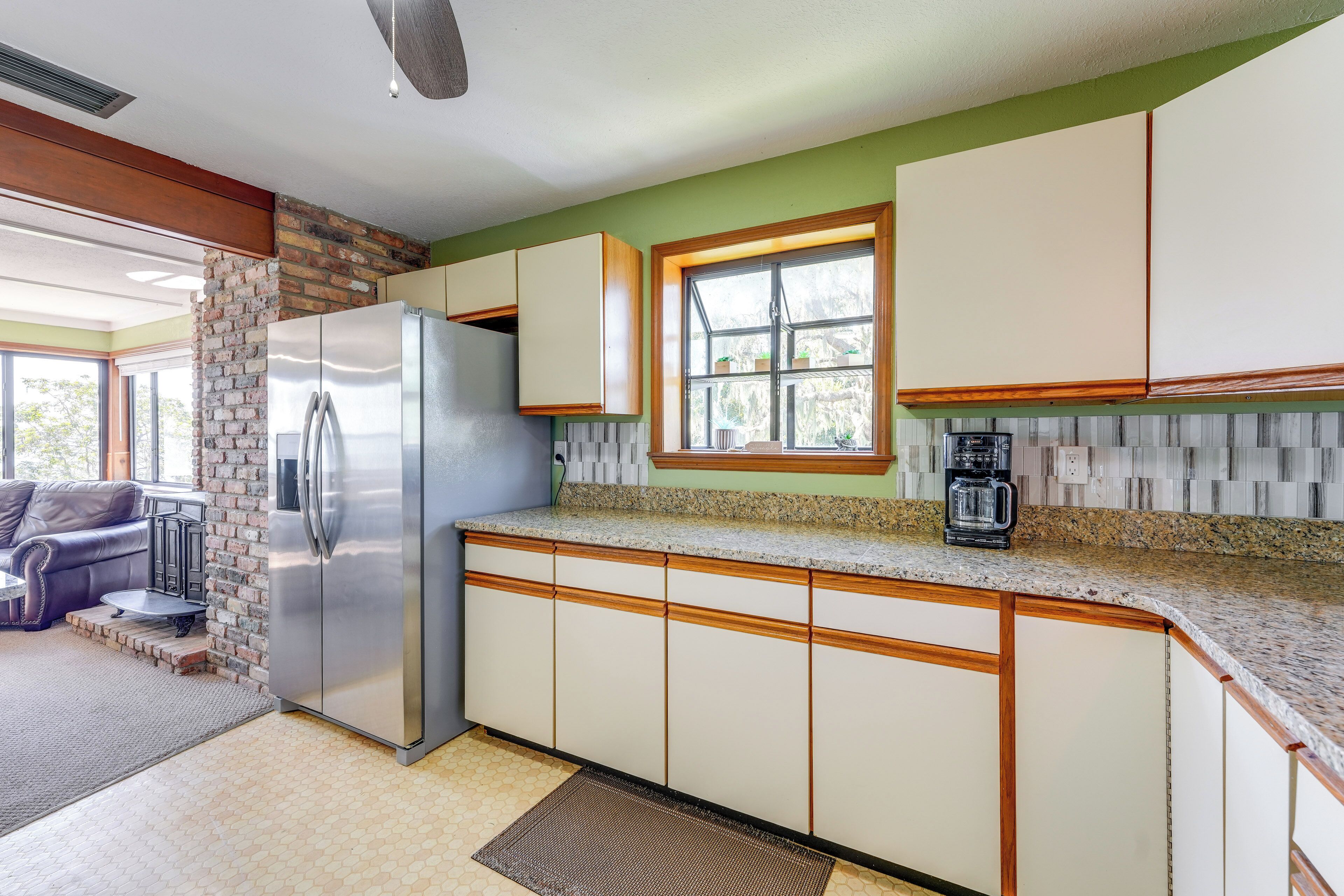 A kitchen with a stainless steel refrigerator, wooden cabinets, a granite countertop, and a window with a view of trees.