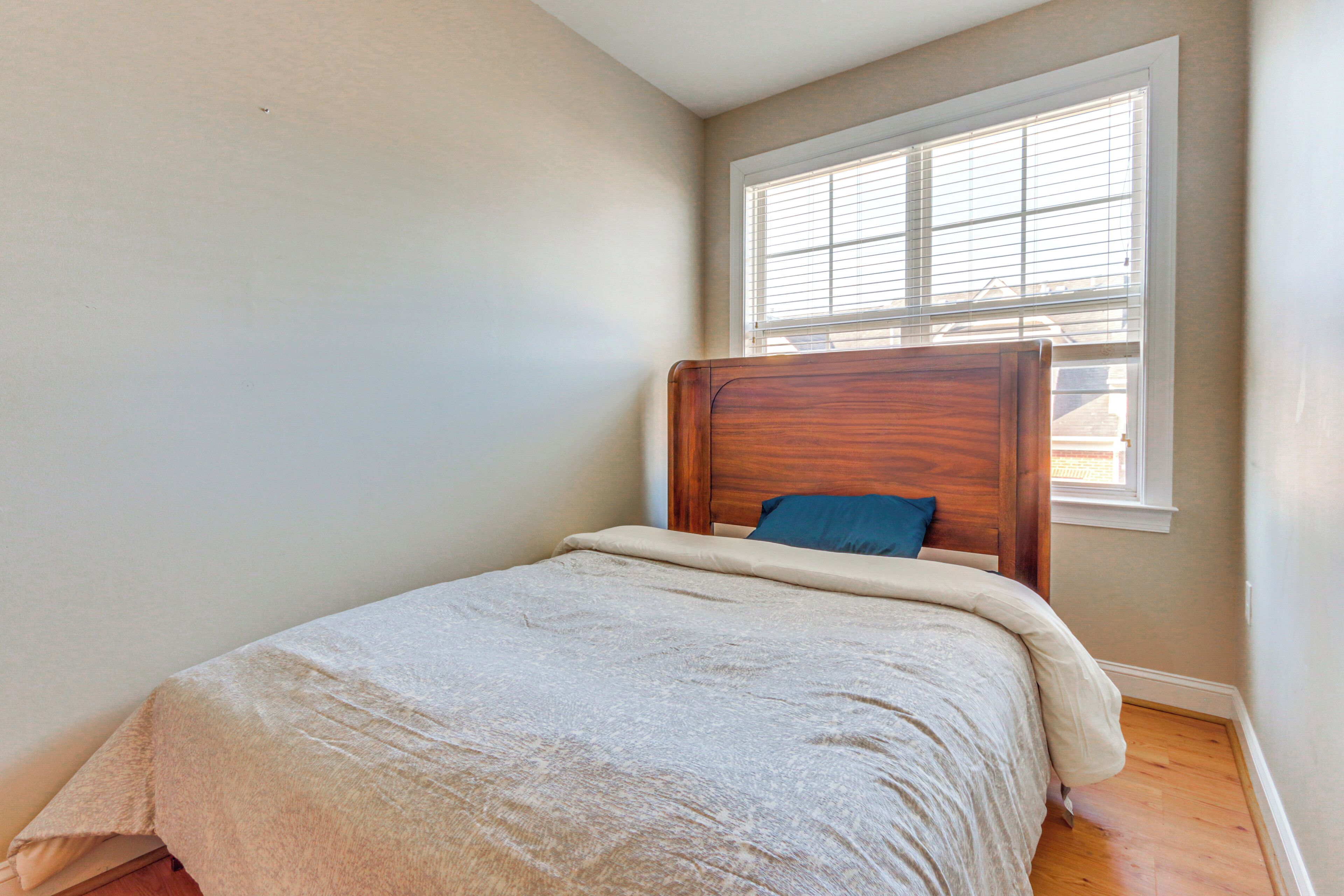 A small, neatly made bed with a wooden headboard and a blue pillow in a room with light-colored walls and a window with blinds.