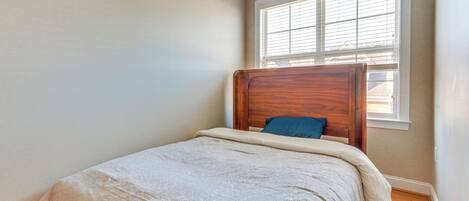 A small, neatly made bed with a wooden headboard and a blue pillow in a room with light-colored walls and a window with blinds.
