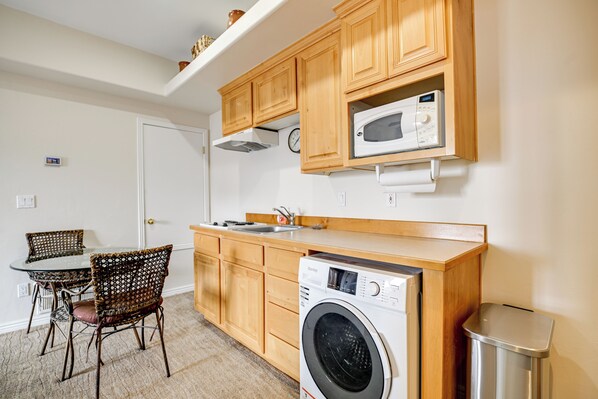 A compact kitchen with wooden cabinets, a microwave, a washing machine, and a dining area with chairs. - Golf Course Views & Private Deck: Mesquite Condo! (Mesquite)