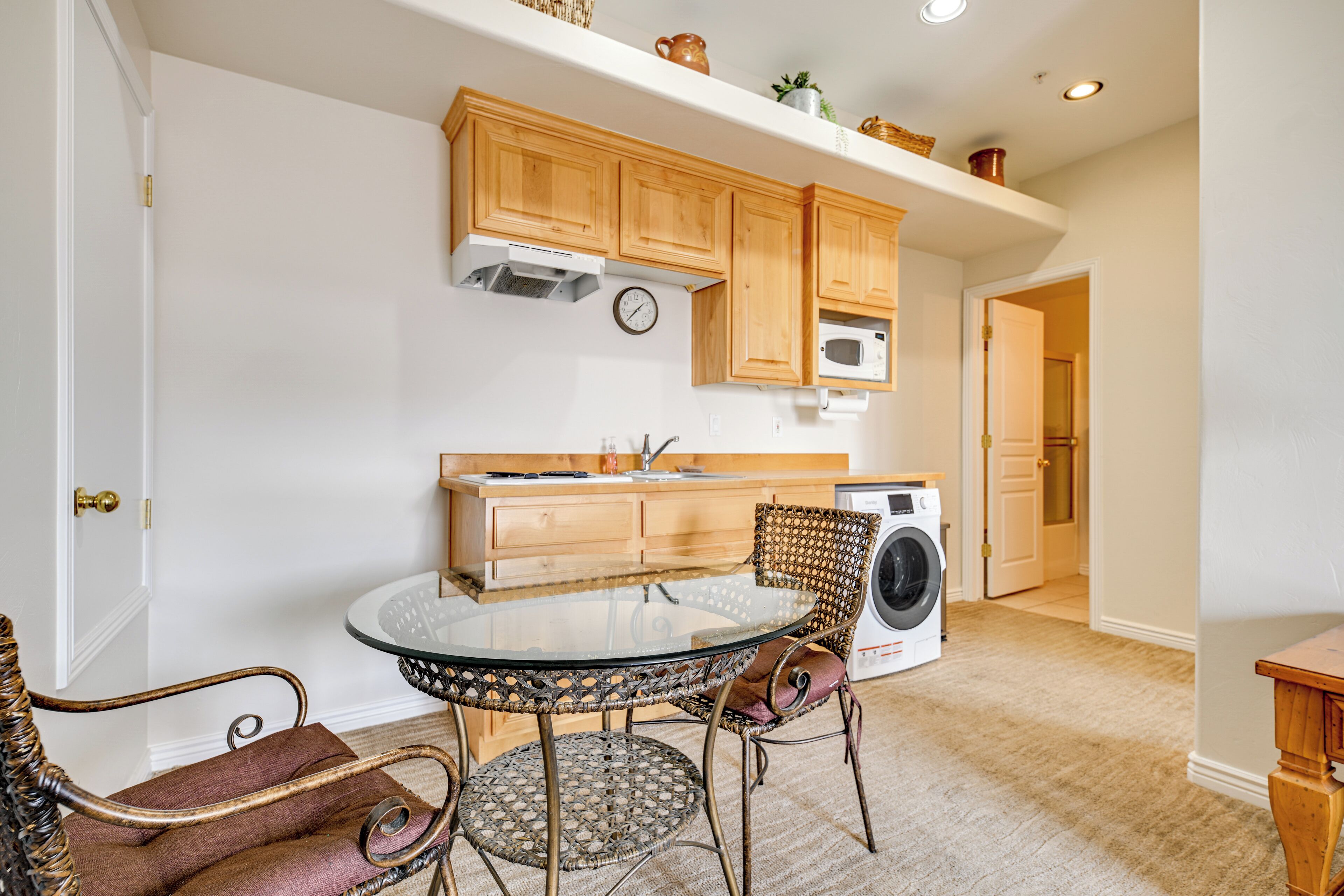 A compact kitchen with wooden cabinets, a microwave, a washing machine, and a round glass table with wicker chairs.
