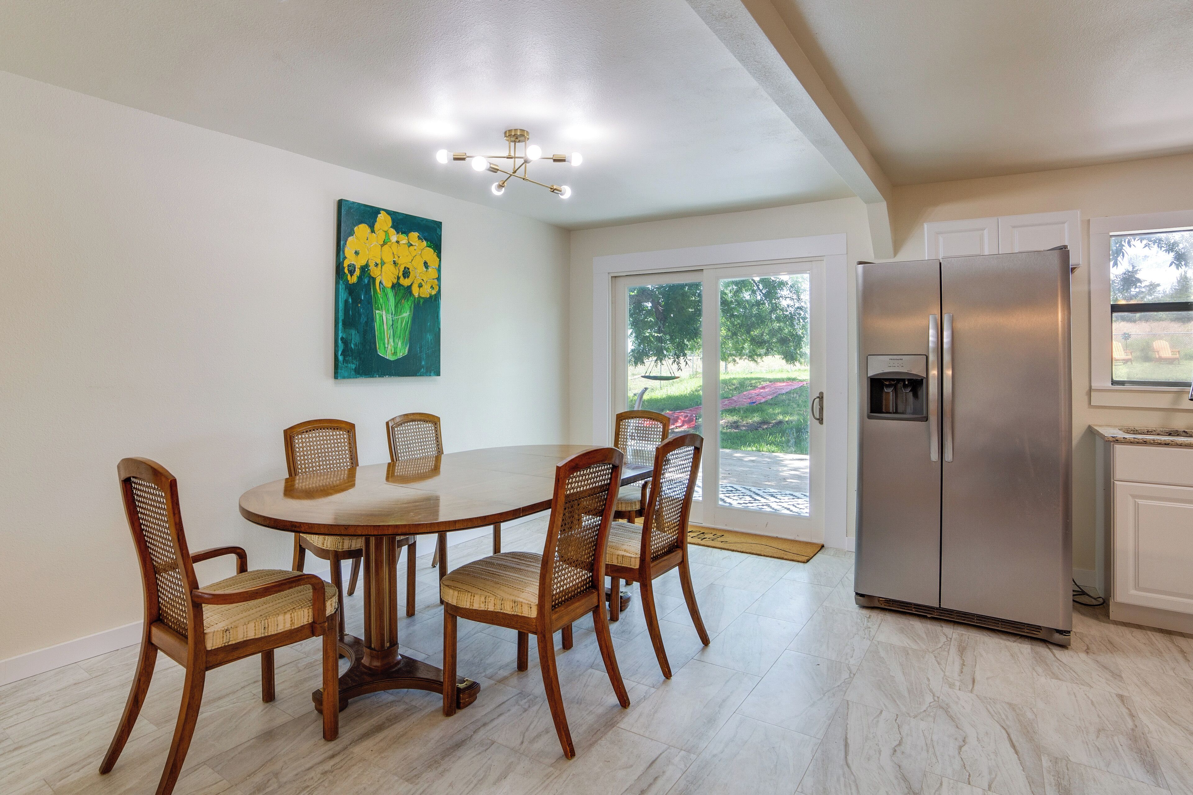 A dining area with a round table and four chairs, a refrigerator, and a painting of yellow flowers on the wall.