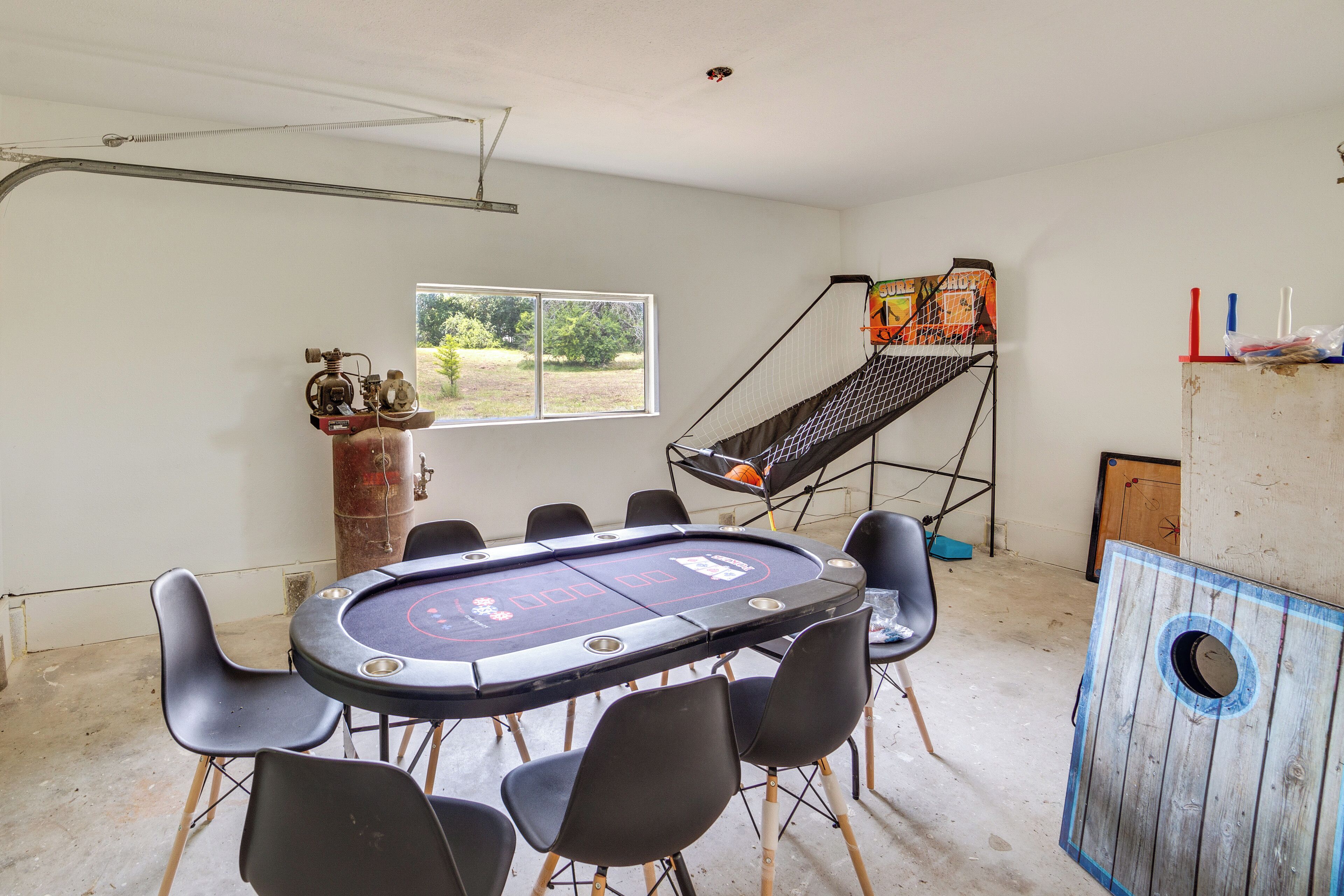 A room with a pool table, chairs, a ladder, and a window with a view of greenery.
