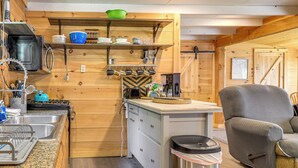 A compact kitchen with wooden walls, a built-in stove, and a sink. There is a trash bin and a chair in the living area.